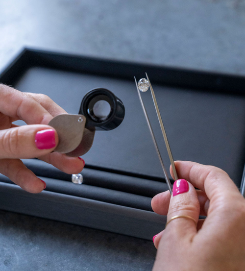 woman with pink nail polish holding diamonds over workstation and inspecting diamond with magnifying glass